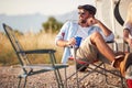 Handsome man with sunglasses and drink sitting in camper chair in front of camper rv, looking into distance and smiling. Outdoors Royalty Free Stock Photo