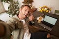 Handsome man takes selfie on his mobile phone with laptop, shows call phone hand sign, smiling at camera, posting photo Royalty Free Stock Photo