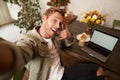 Handsome man takes selfie on his mobile phone with laptop, shows call phone hand sign, smiling at camera, posting photo Royalty Free Stock Photo