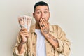 Handsome man with tattoos holding 10 united kingdom pounds banknotes covering mouth with hand, shocked and afraid for mistake