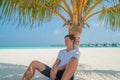 Handsome man wearing white t-shirt and black shorts relaxing under palm tree at tropical sandy beach at island luxury resort Royalty Free Stock Photo