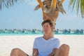 Handsome man in white t-shirt and black shorts relaxing under palm tree at tropical sandy beach at island luxury resort Royalty Free Stock Photo