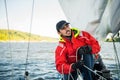 Handsome man on yacht touches white sail called assymetric spinnaker, works with tackle in sea at summer day, under view