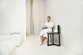 Handsome mature European business man wearing white terry bathrobe, sitting on an armchair and reading magazine while relaxing in Royalty Free Stock Photo