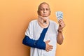 Handsome mature injured man wearing cervical collar and sling holding insurance money puffing cheeks with funny face