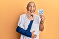 Handsome mature injured man wearing cervical collar and sling holding insurance money sticking tongue out happy with funny