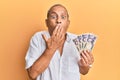 Handsome mature man holding 5000 japanese yen banknotes covering mouth with hand, shocked and afraid for mistake