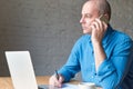 Handsome handsome mature man looks out window and talking on cell phone, sitting at computer, laptop. Man with casual clothes in Royalty Free Stock Photo