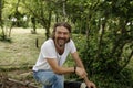 Handsome mature man sitting and laughing in a natural parkland. Bearded and happy man wearing white T-shirt and denim jeans.