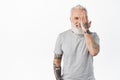 Handsome mature man with tattoos, hiding half of face, cover eye with hand and smiling happy at camera, standing in grey Royalty Free Stock Photo