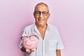 Handsome mature senior man holding piggy bank with glasses looking positive and happy standing and smiling with a confident smile Royalty Free Stock Photo