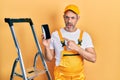 Handsome middle age man with grey hair standing by ladder showing smartphone in shock face, looking skeptical and sarcastic,