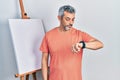 Handsome middle age man with grey hair standing by painter easel stand checking the time on wrist watch, relaxed and confident