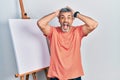 Handsome middle age man with grey hair standing by painter easel stand crazy and scared with hands on head, afraid and surprised Royalty Free Stock Photo