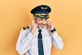 Handsome middle age man with grey hair wearing airplane pilot uniform covering ears with fingers with annoyed expression for the