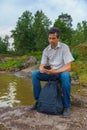 Handsome middle-aged man with smartphone and backpack sitting on bay shore in summer evening. Tourist on the beautiful landscape Royalty Free Stock Photo