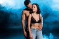 handsome mixed race man hugging attractive girl in lace black bra on black with blue smoke. Royalty Free Stock Photo