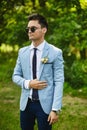 Handsome model man wearing in the fashionable blue jacket with a narrow tie and stylish sunglasses stands in the green Royalty Free Stock Photo