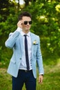 Handsome model man wearing in the fashionable blue jacket with a narrow tie and stylish sunglasses stands in the green Royalty Free Stock Photo