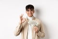 Handsome modern man in glasses showing dollar bills and okay gesture, making money, standing with cash on white Royalty Free Stock Photo