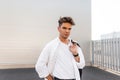 Handsome modern young man in fashionable white clothes with stylish black cloth bag with trendy hairstyle stands near a white Royalty Free Stock Photo