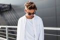 Handsome modern young man in fashionable white clothes in stylish black sunglasses with trendy hairstyle stands on a bright sunny Royalty Free Stock Photo