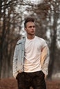Handsome modern young man in a vintage white T-shirt in a blue denim jacket with a fashionable hairstyle posing outdoors in a park Royalty Free Stock Photo