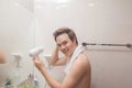Handsome naked young man drying hair with hairdryer, looking at mirror at home