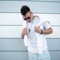 Handsome nice young hipster man in trendy white t-shirt in stylish sunglasses with fashionable hairstyle posing near a modern Royalty Free Stock Photo
