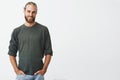 Handsome nordic man with beard and stylish hairstyle in grey shirt and jeans smiling, looking in camera, keeps hands in