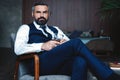 Handsome pensive man is touching his beard, looking away and thinking while sitting in armchair indoors.