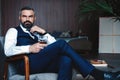 Handsome pensive man is touching his beard, looking away and thinking while sitting in armchair indoors.