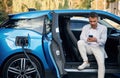 Handsome person with smartphone. Man with blue electric car on the charge station