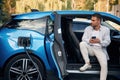 Handsome person with smartphone. Man with blue electric car on the charge station