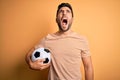 Handsome player man with beard playing soccer holding footballl ball over yellow background angry and mad screaming frustrated and Royalty Free Stock Photo