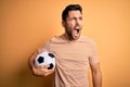 Handsome player man with beard playing soccer holding footballl ball over yellow background angry and mad screaming frustrated and Royalty Free Stock Photo