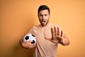 Handsome player man with beard playing soccer holding footballl ball over yellow background doing stop gesture with hands palms, Royalty Free Stock Photo