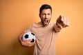 Handsome player man with beard playing soccer holding footballl ball over yellow background pointing displeased and frustrated to Royalty Free Stock Photo