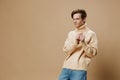 a handsome, pleasant, smiling young man in a long beige sweater stands against a plain background and holds his sleeves