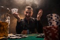 Handsome poker player with two aces in his hands and chips sitting at poker table in a dark room full of cigarette smoke