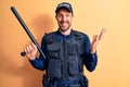 Handsome policeman wearing uniform and bulletprof holding baton over yellow background celebrating achievement with happy smile