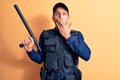 Handsome policeman wearing uniform and bulletprof holding baton over yellow background covering mouth with hand, shocked and