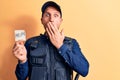 Handsome policeman wearing uniform and bulletprof holding reminder with help message covering mouth with hand, shocked and afraid