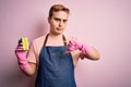 Handsome redhead man doing housework wearing apron and gloves using cleaner scourer with angry face, negative sign showing dislike