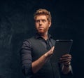 Portrait of a redhead man in formal wear holding a tablet in studio against a dark textured wall