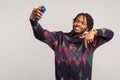 Handsome self confident african male blogger with dreadlocks showing subscribe gesture looking at camera of smartphone with toothy