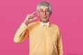 Handsome senior man in bright yellow shirt raises right hand up and shows quantity of tea in his cup with fingers. Old man stands Royalty Free Stock Photo