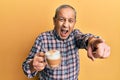 Handsome senior man with grey hair drinking a cup coffee pointing displeased and frustrated to the camera, angry and furious with Royalty Free Stock Photo