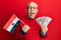 Handsome senior man with grey hair holding egypt flag and egyptian pounds banknotes sticking tongue out happy with funny Royalty Free Stock Photo