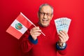 Handsome senior man with grey hair holding peru flag and peruvian sol banknotes winking looking at the camera with sexy